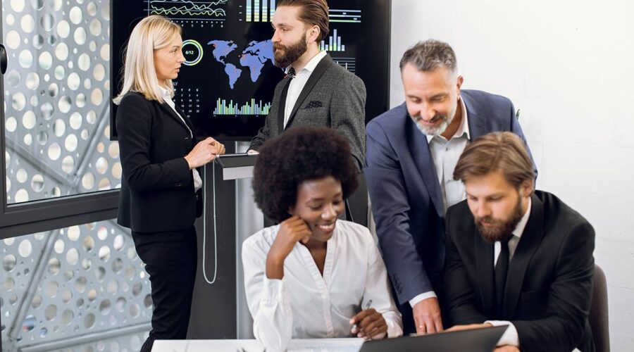 Positive purposeful multiracial business colleagues, working together over joint business project. Focus on mature blond woman and bearded man behind, using interactive screen in modern meeting room